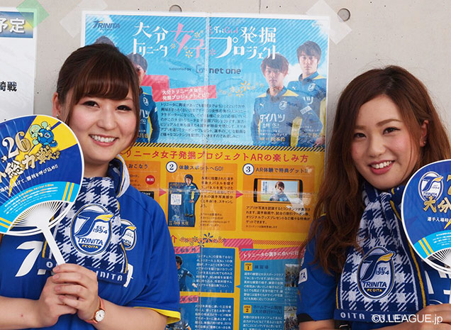 【明治安田Ｊ２ 第26節 大分vs群馬】「大分トリニータ女子発掘プロジェクト」ポスターを背景に笑みを見せる大分の女性ファン・サポーター（1/4）